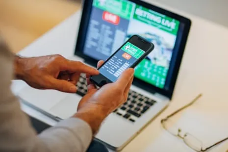 Un usuario consulta cuotas de apuestas en su teléfono móvil frente a un ordenador, imagen que ilustra el mercado del juego en línea no regulado.